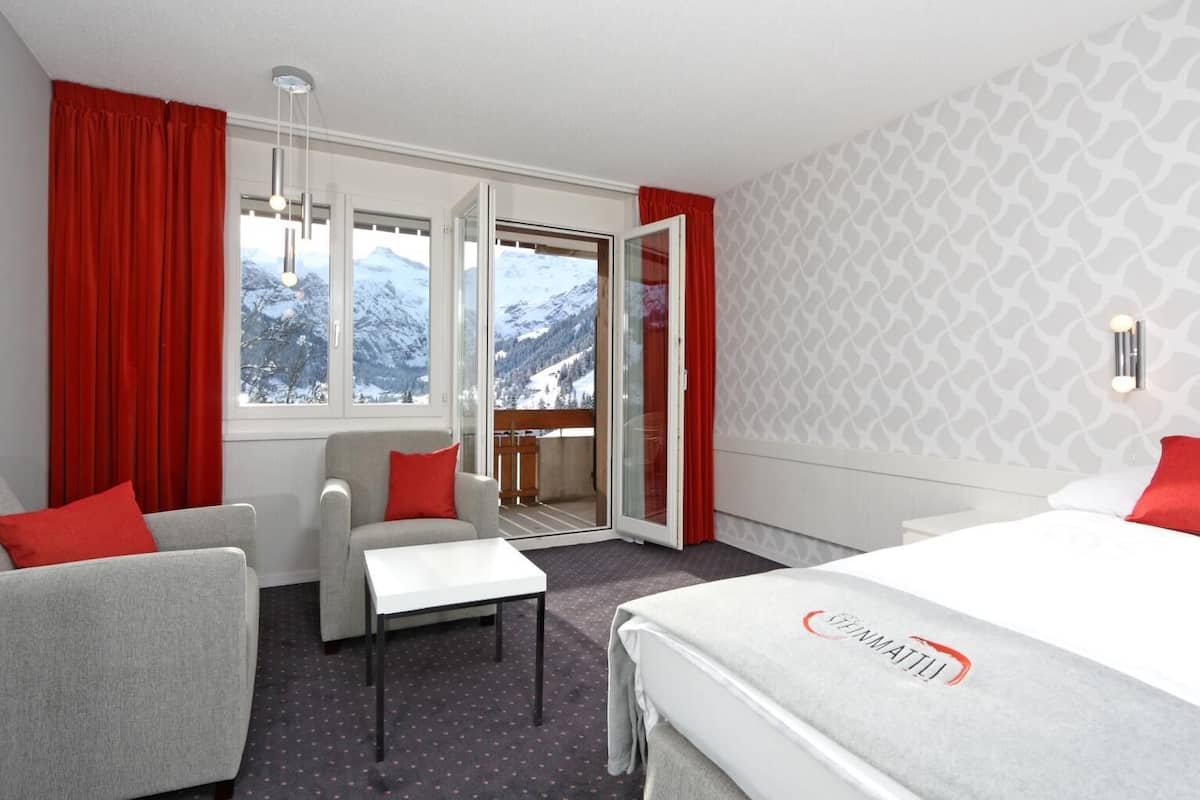 A hotel room with a bed, two armchairs, a small table, and a balcony with a mountain view.
