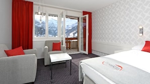 A hotel room with a bed, two armchairs, a small table, and a balcony with a mountain view.