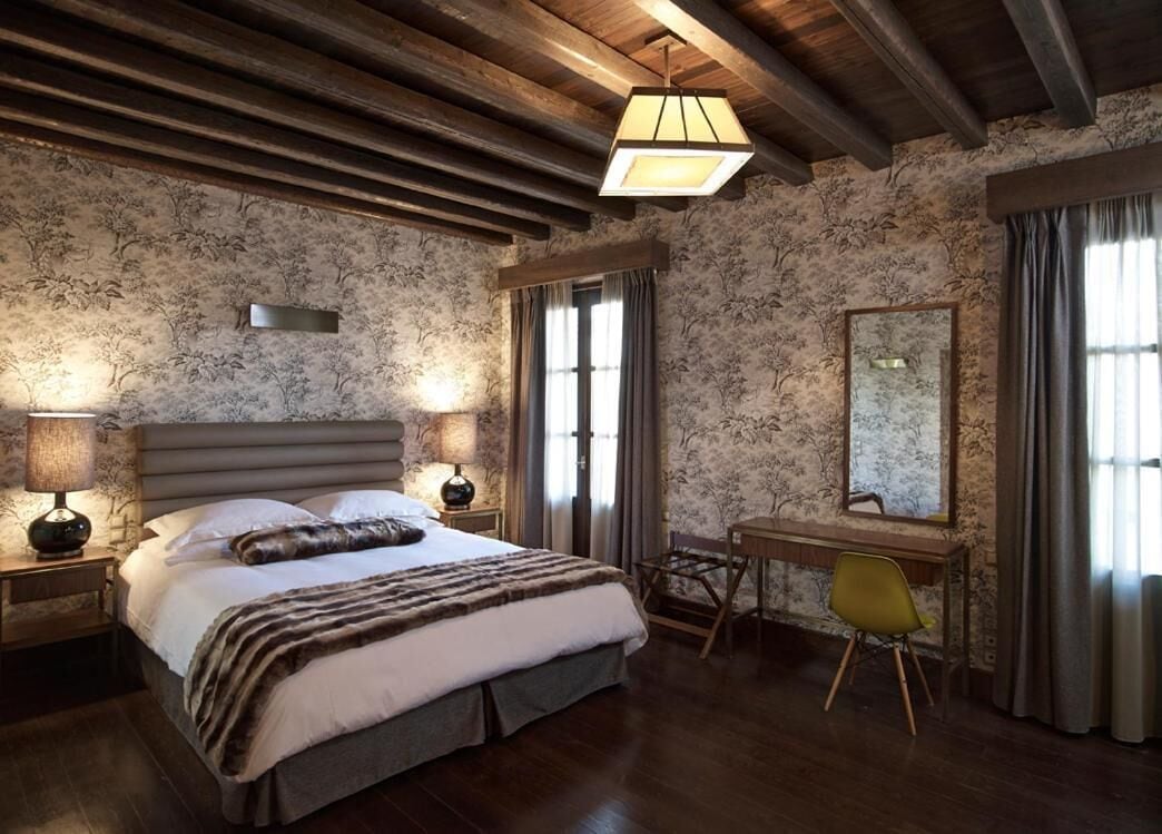 A bedroom with a large bed, a desk, and a chair. There is a window with curtains and a lamp on the bedside table.