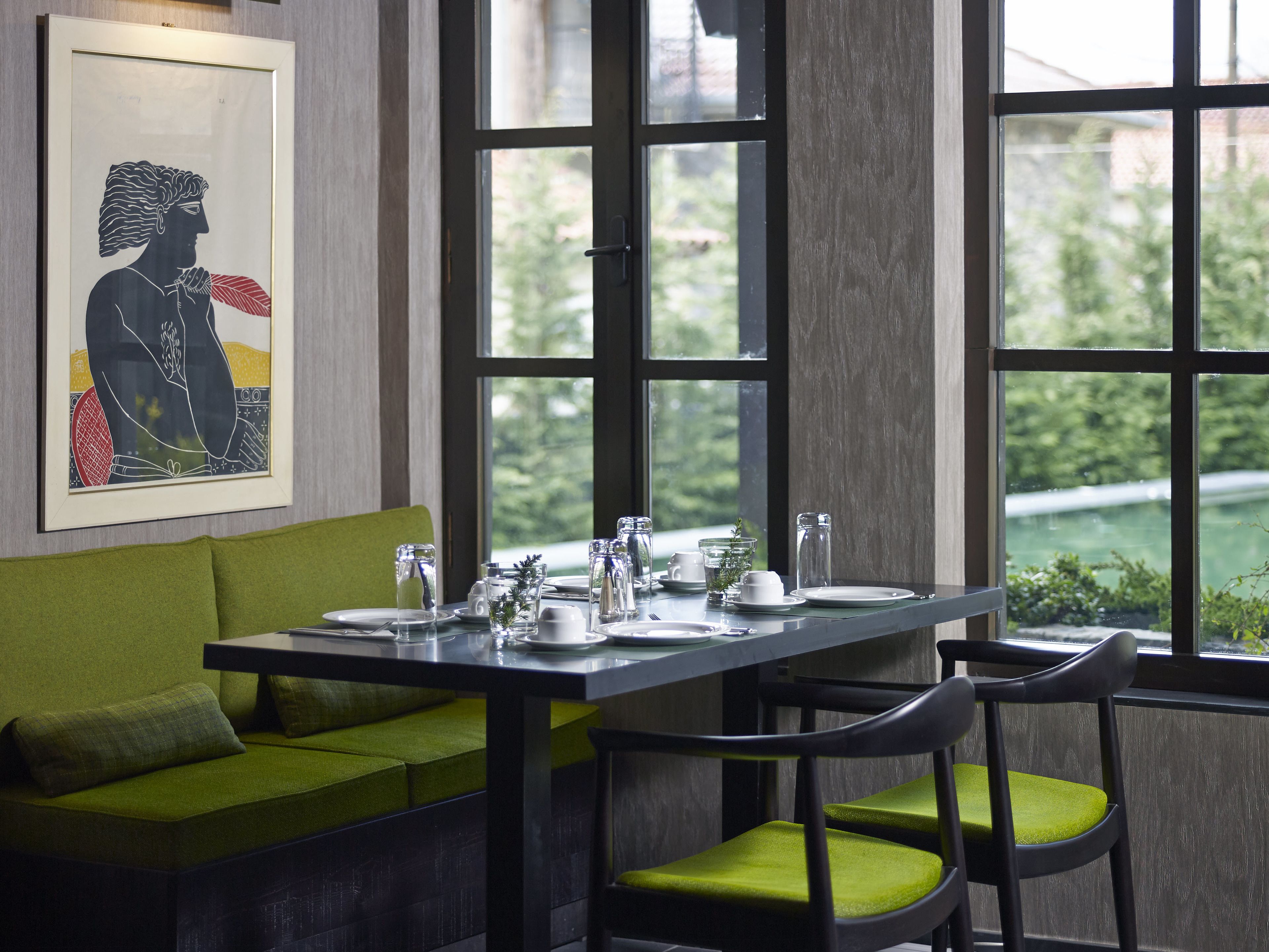 A dining area with a green sofa, two black chairs with green cushions, a table set with plates, glasses, and cutlery, and a framed artwork on the wall.