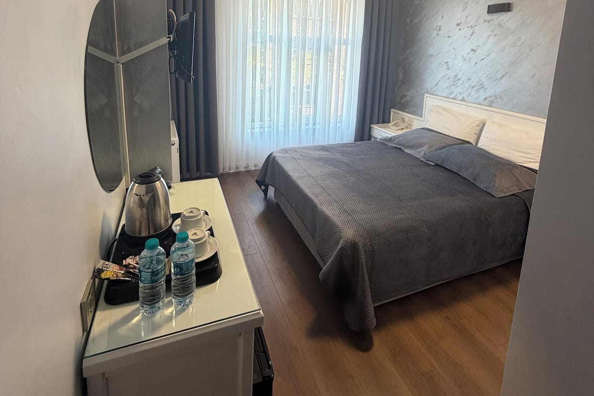 A modern hotel room with a bed, bedside table, and a desk with a kettle and water bottles.