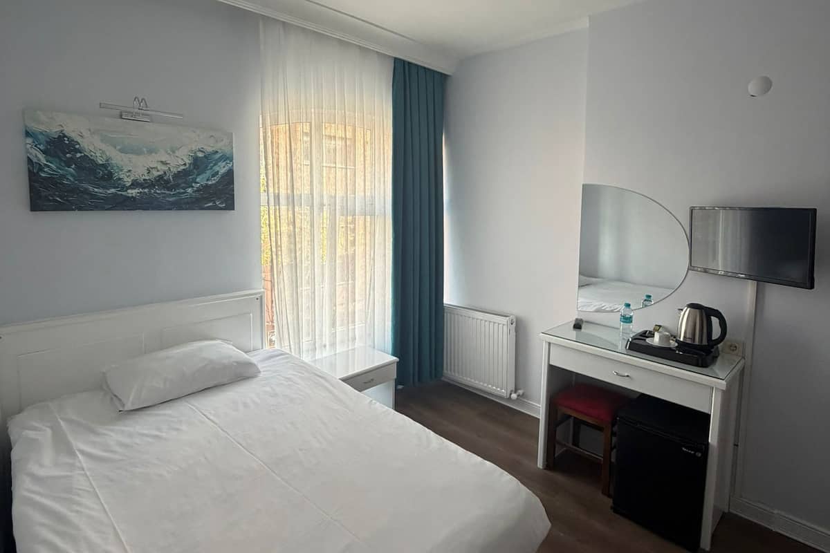 A hotel room with a bed, a desk, a chair, a radiator, a window with curtains, and a painting on the wall.