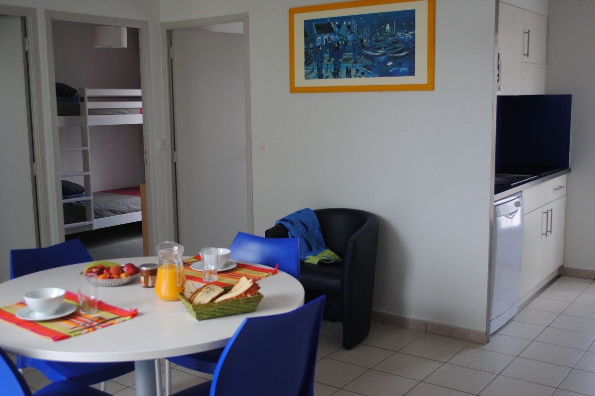 Un coin repas avec une table blanche, des chaises bleues, un lit superposé, une cuisine avec un téléviseur et un tableau au mur.
