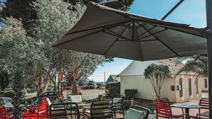 Un espacio al aire libre con sillas y mesas bajo un gran paraguas, rodeado de árboles y un edificio al fondo.
