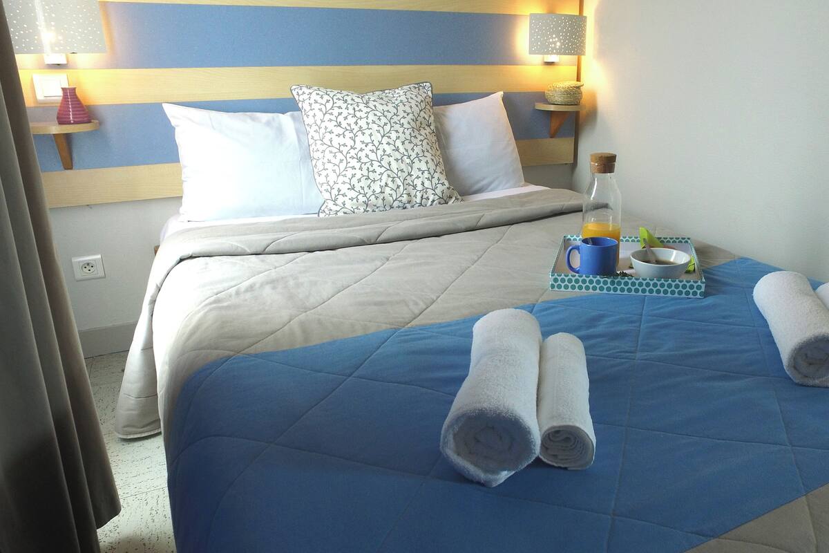 Habitación de hotel con cama, almohadas, mesita de noche con lámpara, una botella, un cuenco y toallas enrolladas.