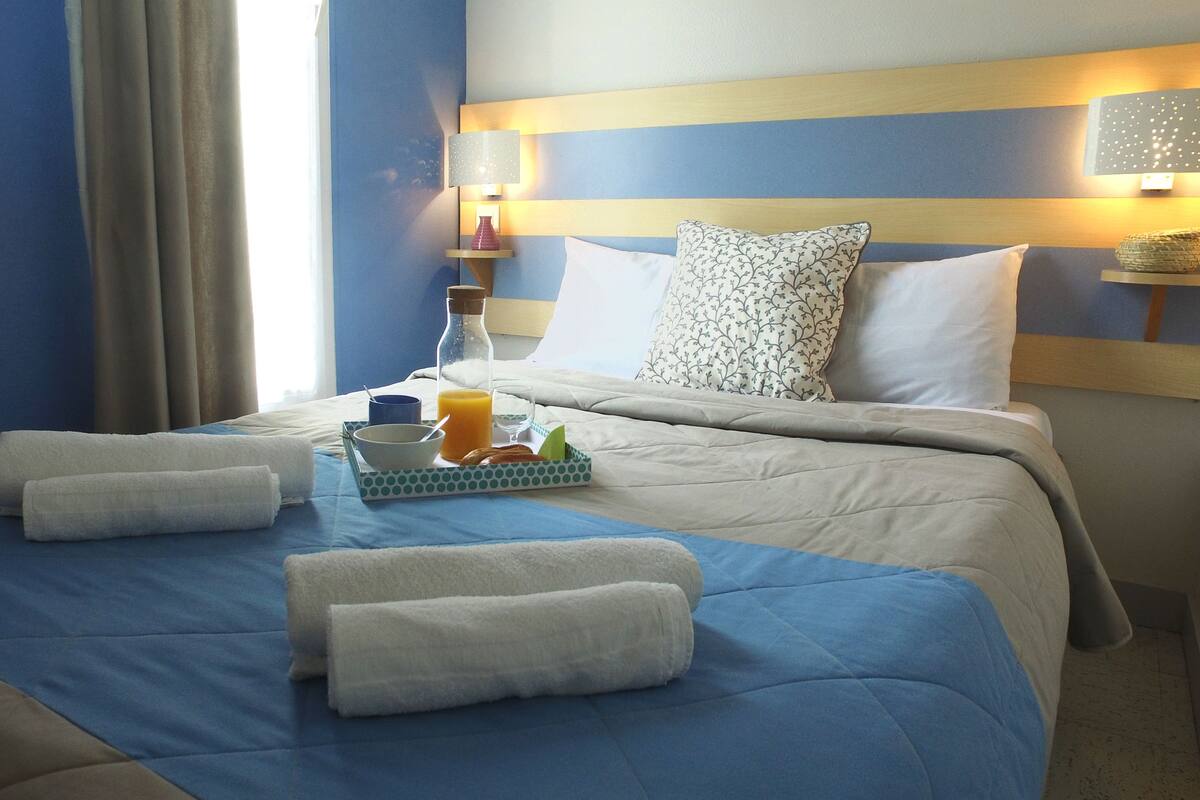 Habitación de hotel con una cama bien hecha, una mesita de noche con lámpara, un jarrón y una bandeja con alimentos para el desayuno.