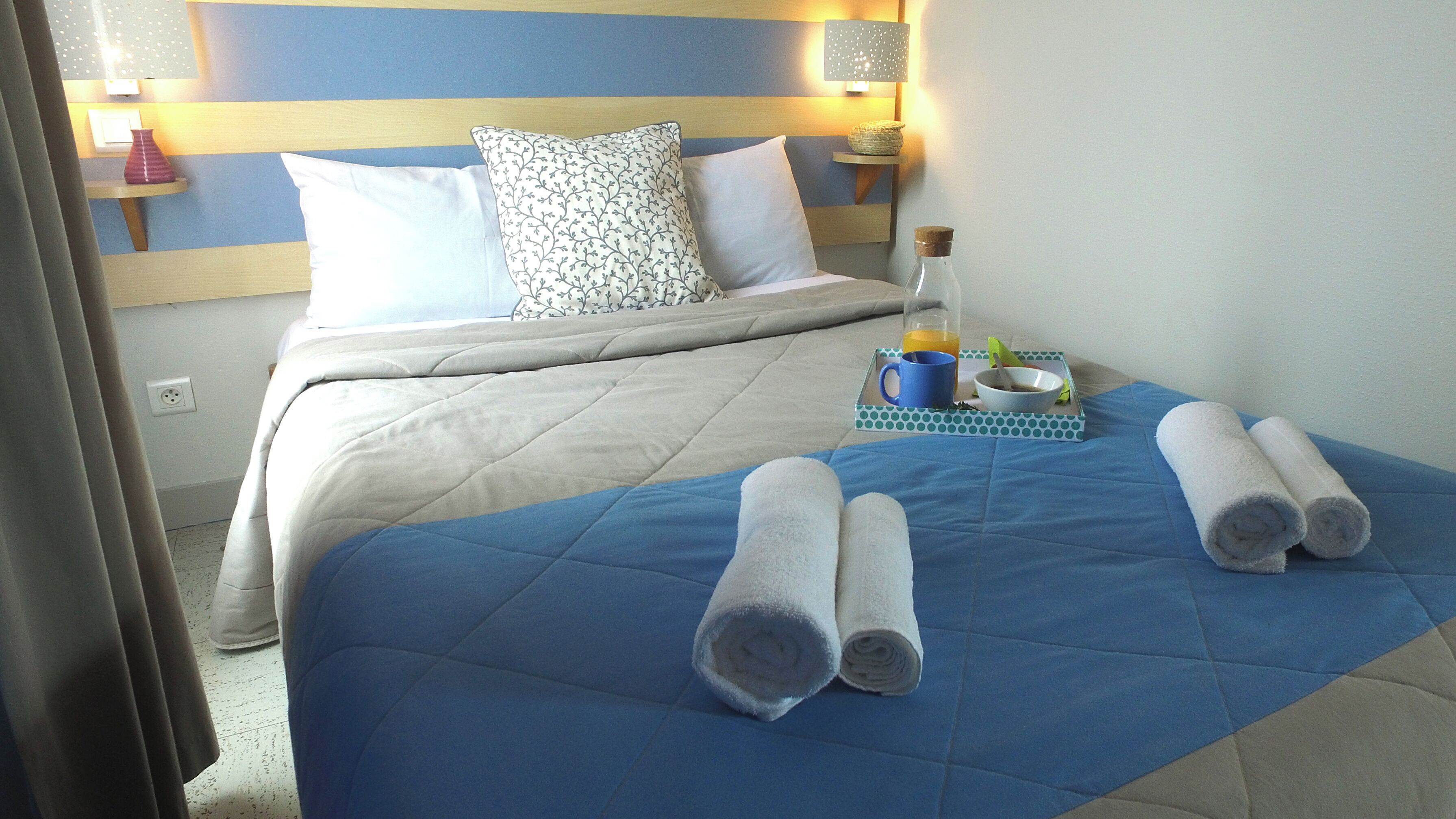 A hotel room with a bed, pillows, a bedside table with a lamp, a bottle, a bowl, and rolled towels.