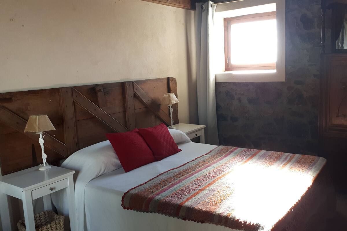 A bedroom with a wooden headboard, a bed with white bedding and red pillows, a bedside table with a lamp, and a window with sunlight.