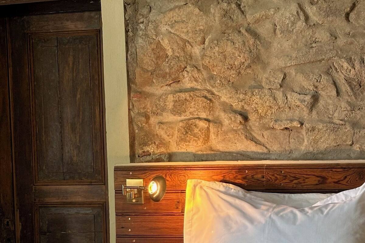 A bed with white linens and a rolled towel, a wooden nightstand with a lamp, and a stone wall.
