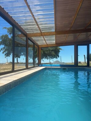 A swimming pool with a glass ceiling and wooden beams, surrounded by trees and a clear sky.