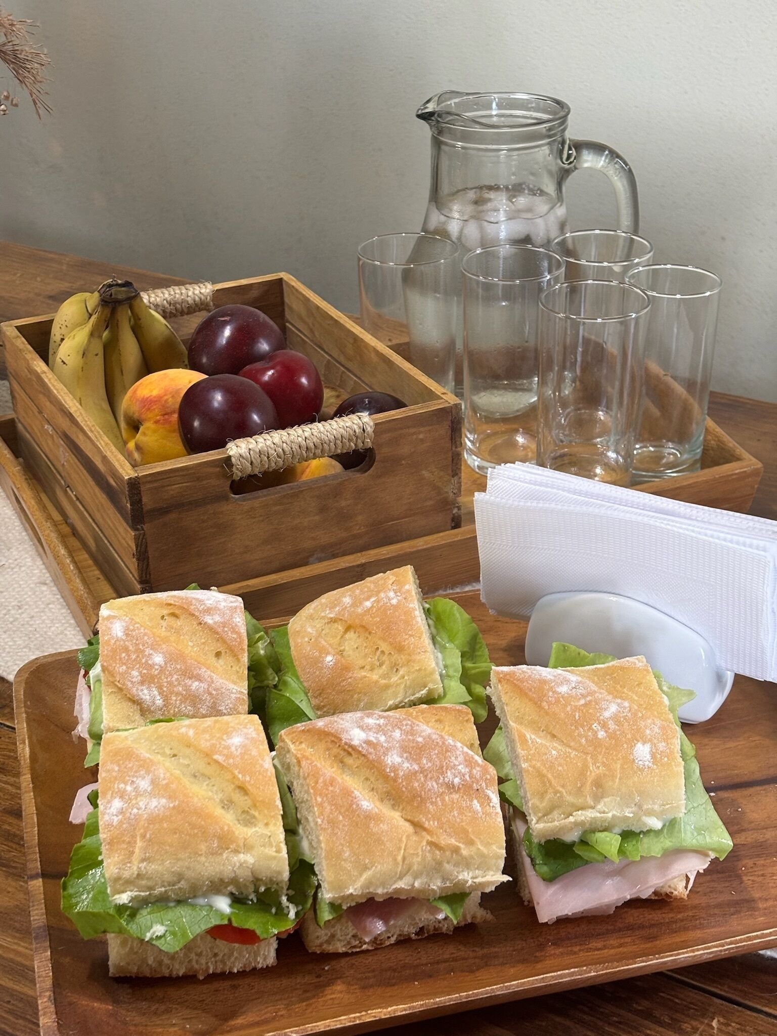 Un plateau en bois avec des sandwichs, un panier de fruits et des verres transparents.