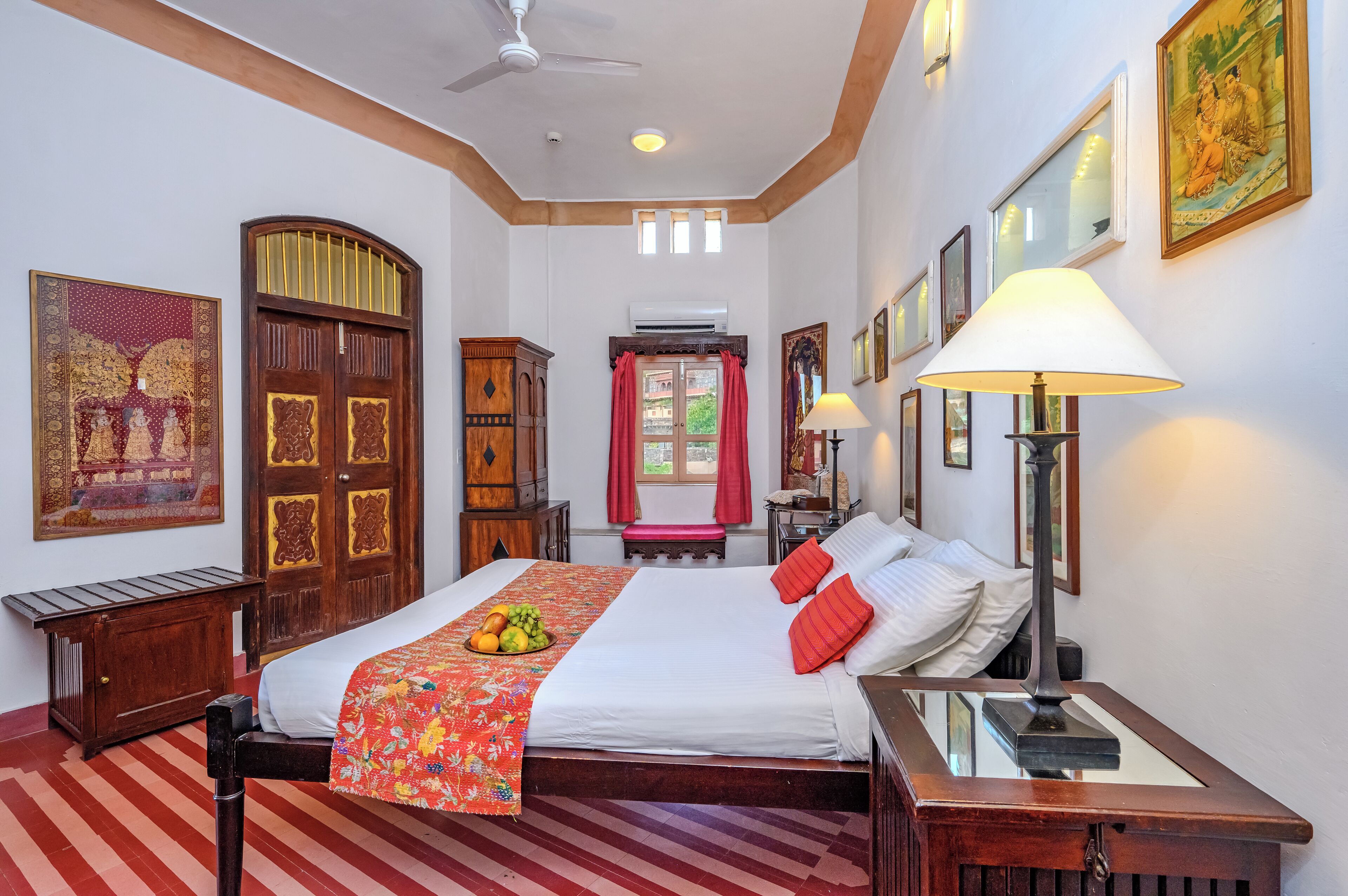 A hotel room with a large bed, a wooden headboard, a bedside table, a lamp, and a window with red curtains.