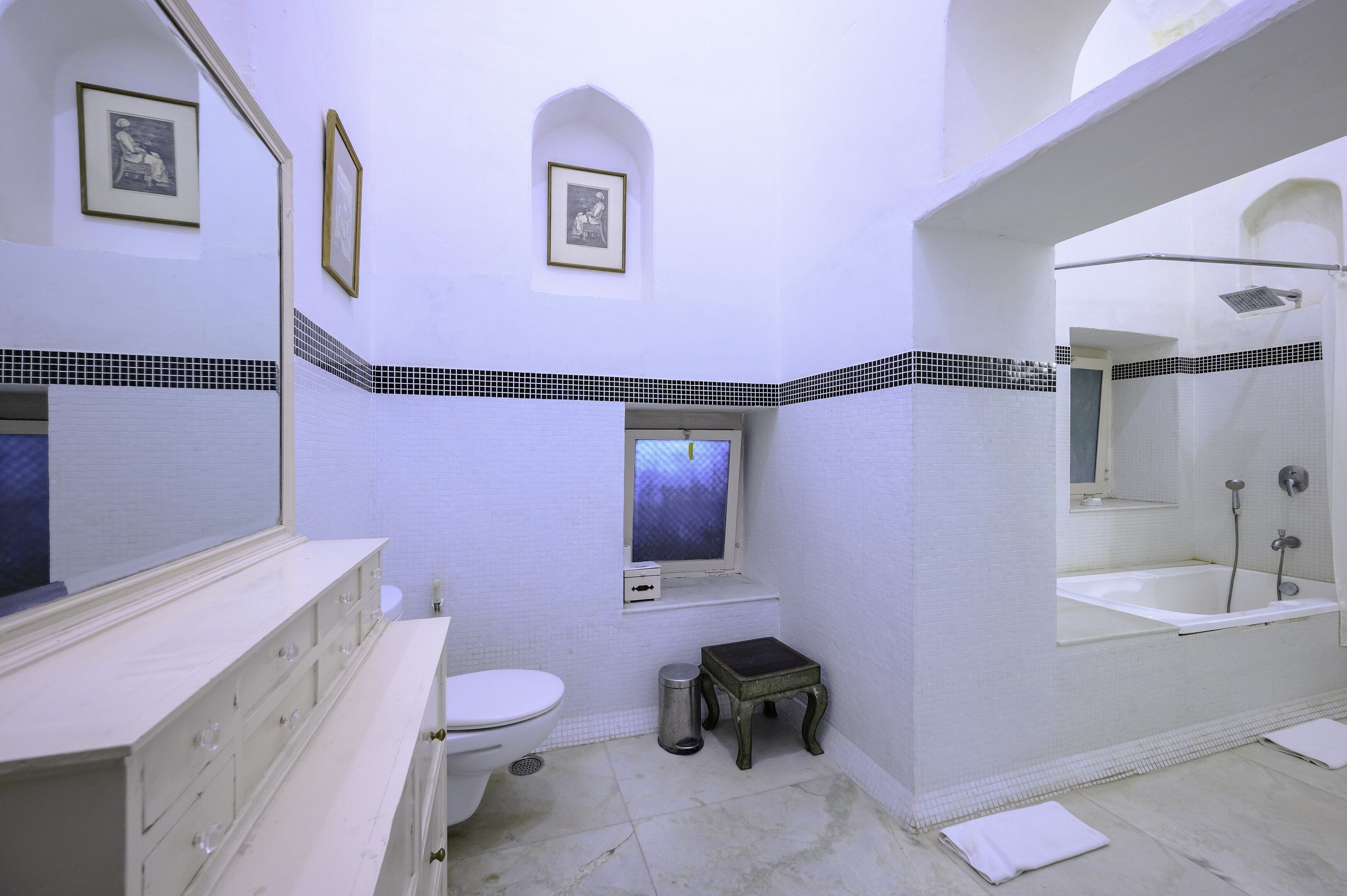 Une salle de bain avec un meuble-lavabo blanc, des toilettes, un petit tabouret, une fenêtre et une baignoire avec douche.