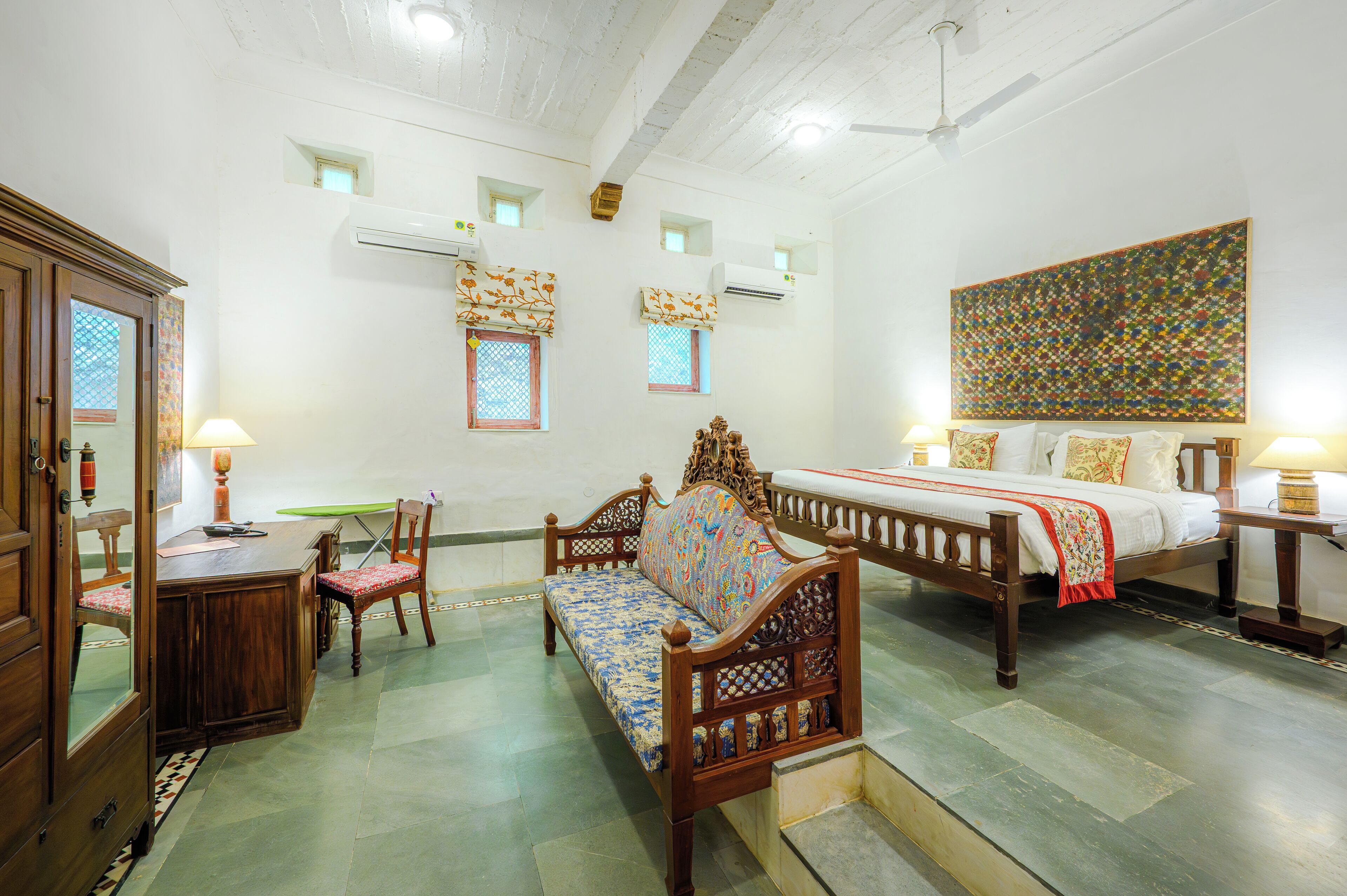 A traditional room with a wooden bed, a sofa, a dining table, and chairs. There are air conditioning units on the walls and a ceiling fan.