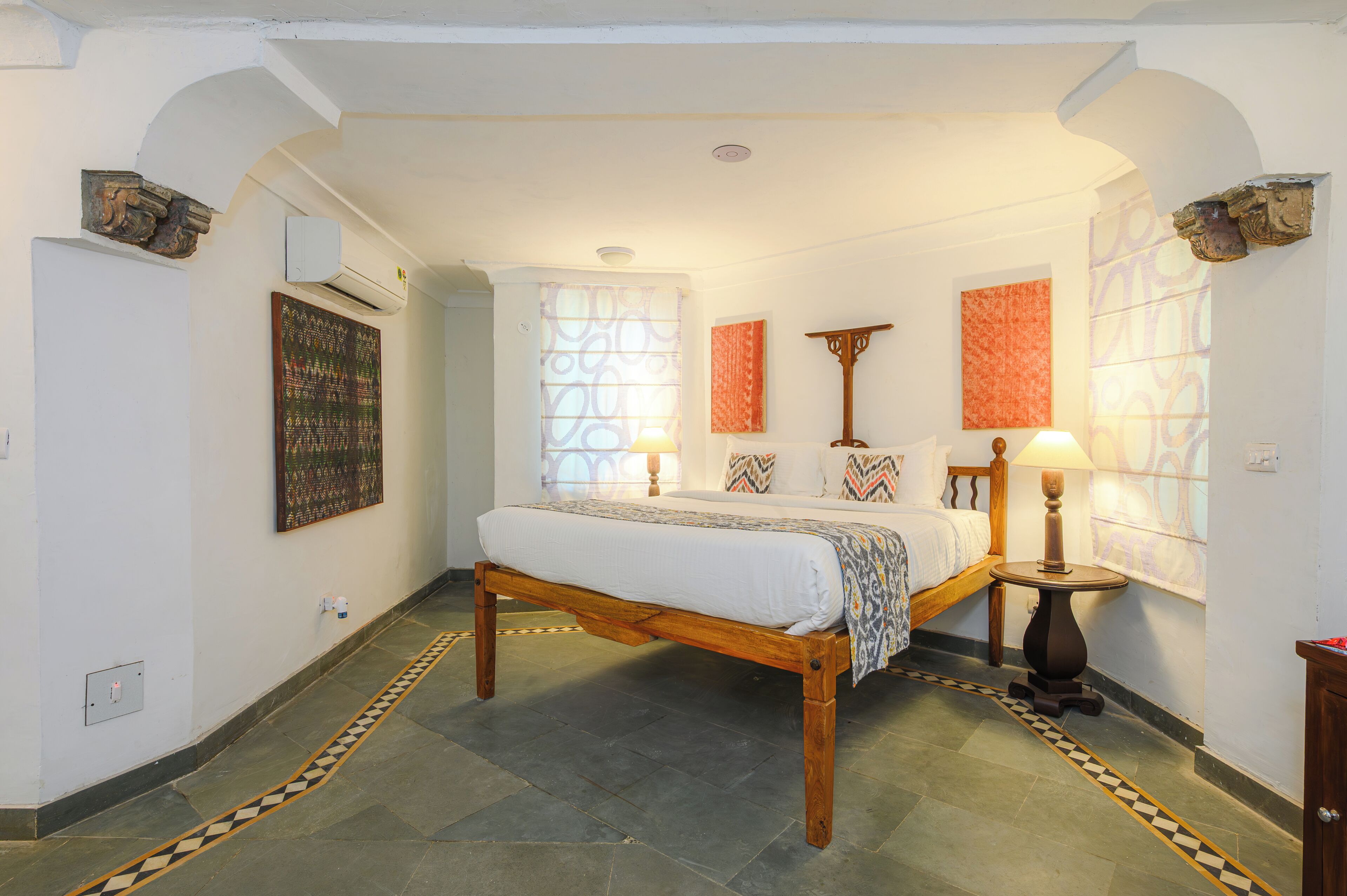 A bedroom with a wooden bed, two bedside lamps, a window with patterned curtains, and a wall-mounted air conditioner.