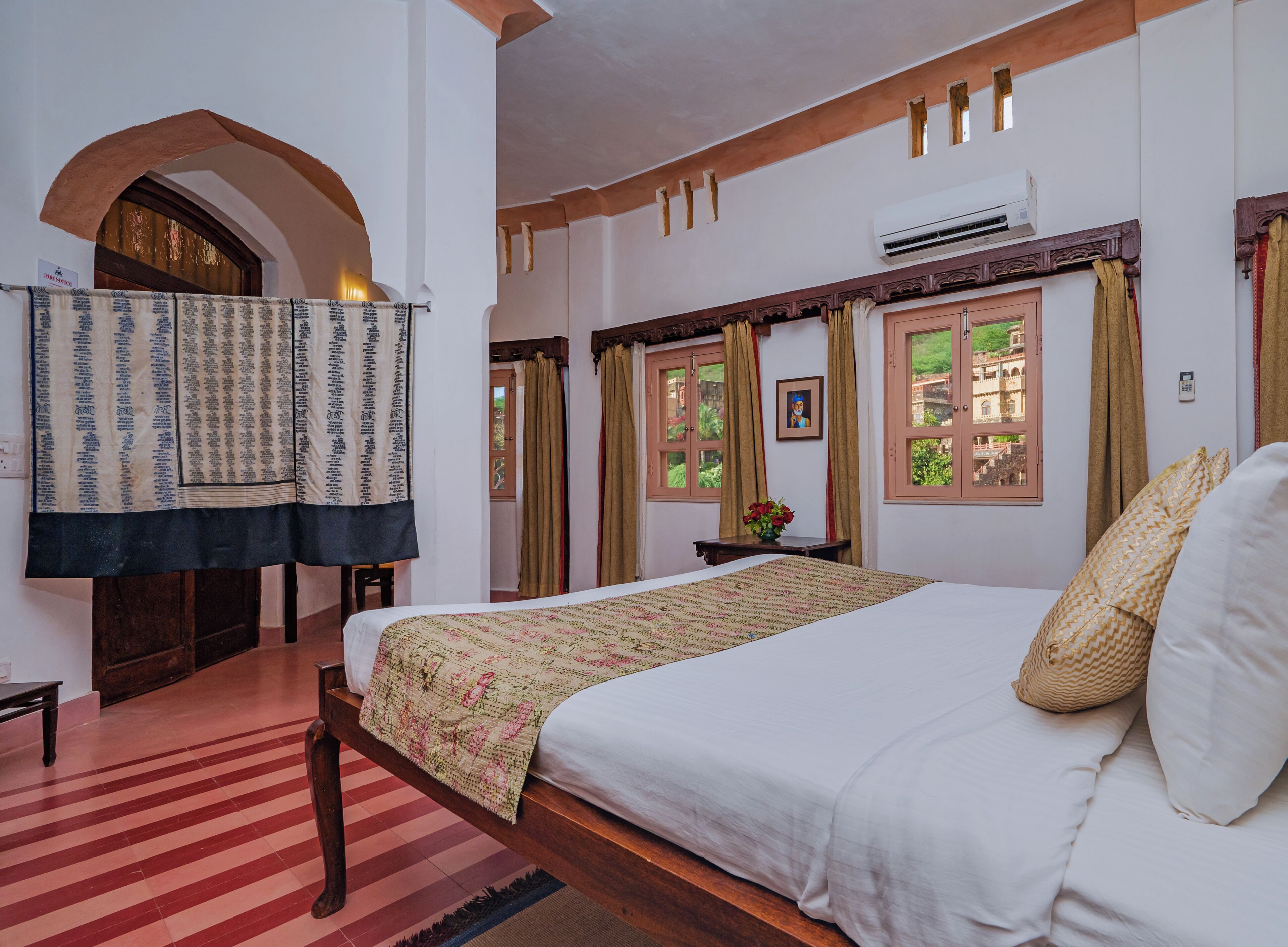 A hotel room with a large bed, a wooden bench, a small table with a vase, and a view of greenery outside.