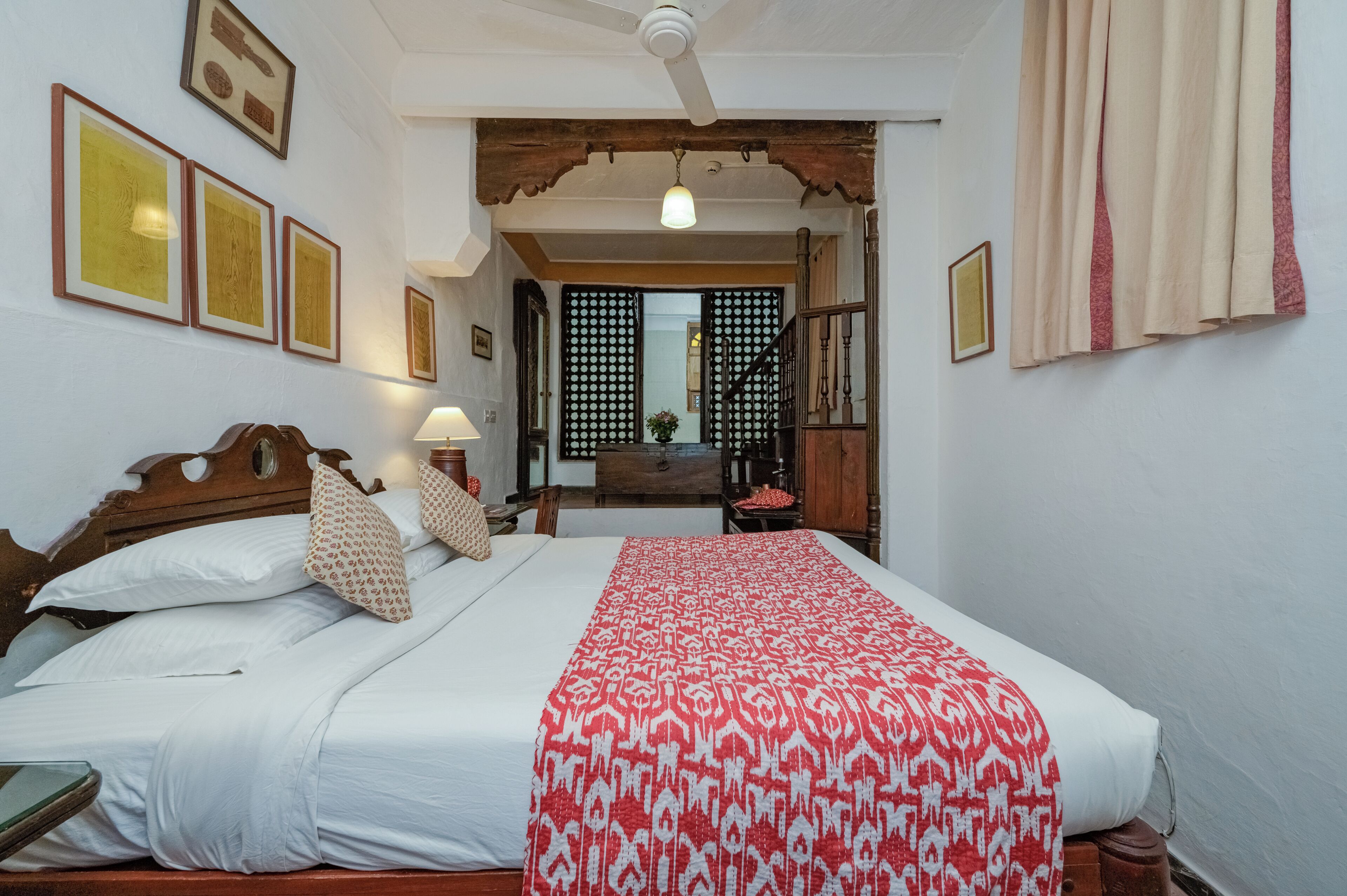 A bedroom with a wooden bed, white bedding, a red patterned bedspread, a nightstand with a lamp, and a view of a balcony through a wooden door.