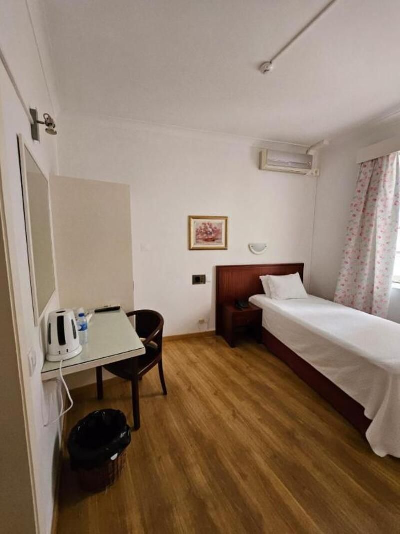 Una habitación pequeña y funcional con una cama individual, un escritorio con hervidor, una silla y un cesto de basura.
