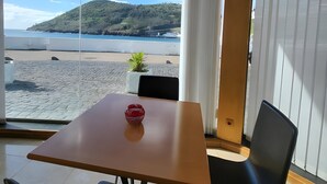 A room with a table and chairs, a view of a beach and a hill, and a window with curtains.