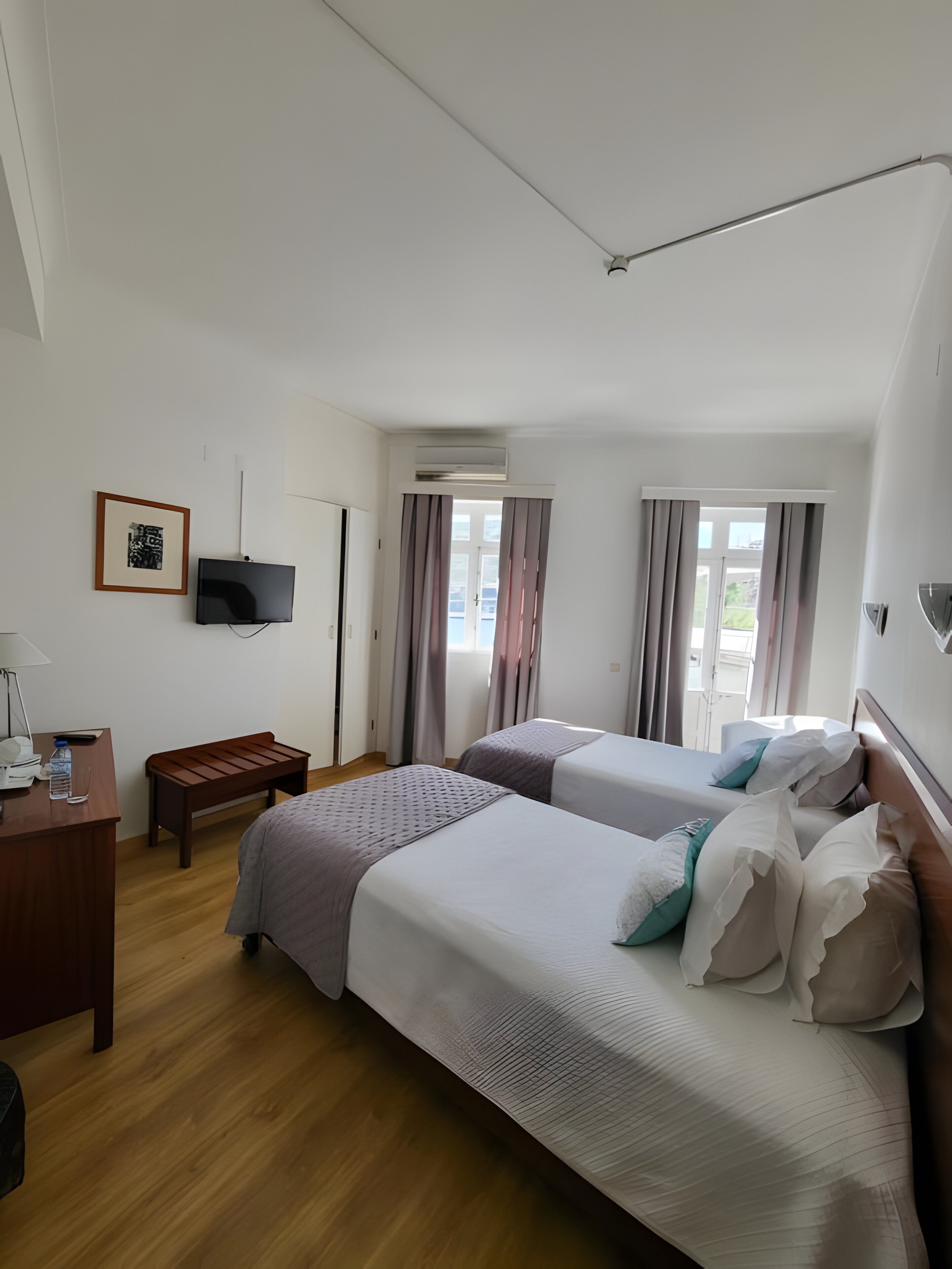 A hotel room with two beds, a desk with a telephone and water bottle, a TV mounted on the wall, and an air conditioning unit.