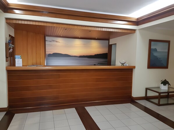 A reception desk with a wooden finish, a framed landscape painting, and a small table with a plant. - Hotel Beira Mar (Angra do Heroismo)
