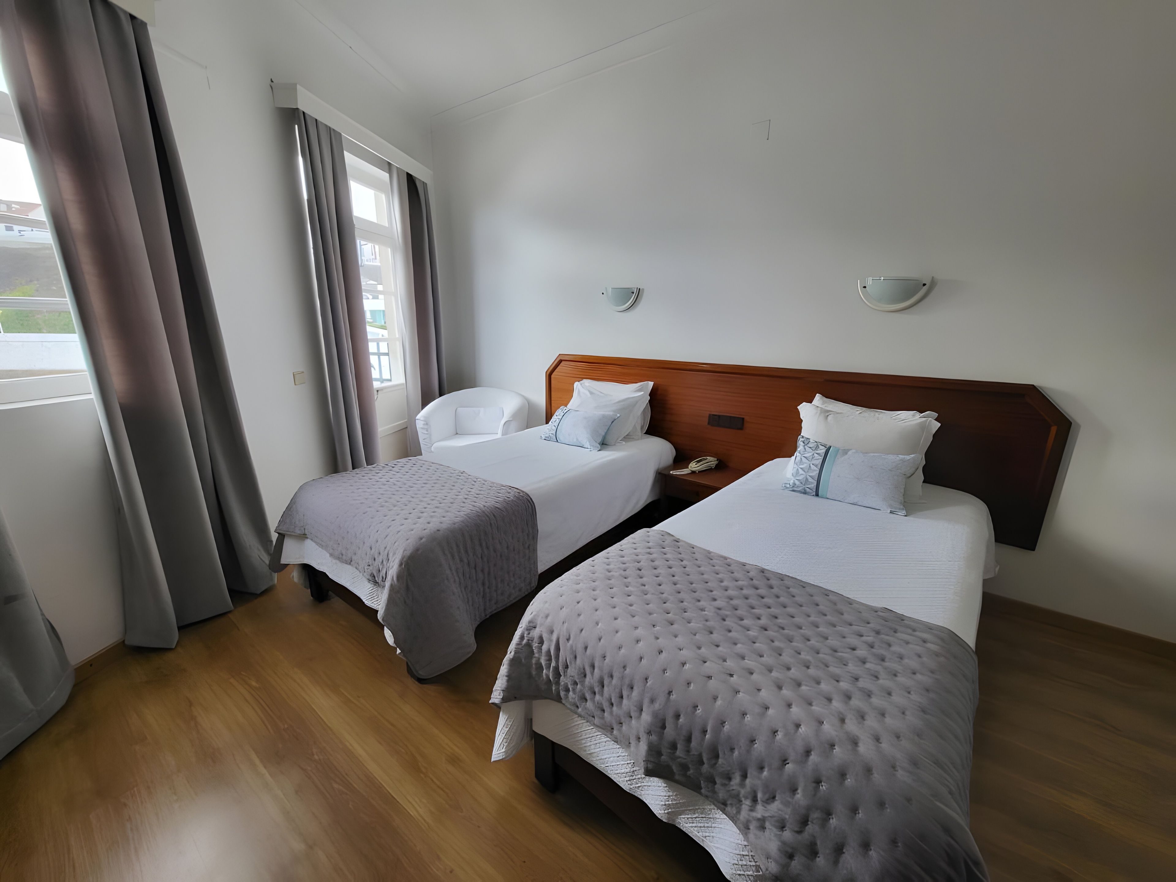 A hotel room with two beds, a chair, a bedside table with a phone, and a window with curtains.