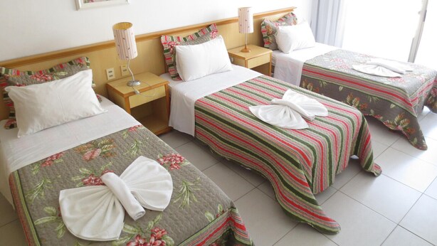 Quarto de hotel com três camas, cada uma com colcha florida e toalha sobre a cama. Há mesinhas de cabeceira com abajures e uma janela com cortinas.