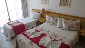 Quarto de hotel com duas camas, cada uma com colcha florida e toalhas brancas dobradas em laço. Há duas imagens florais emolduradas na parede, uma mesa de cabeceira com abajur e uma janela com cortinas.