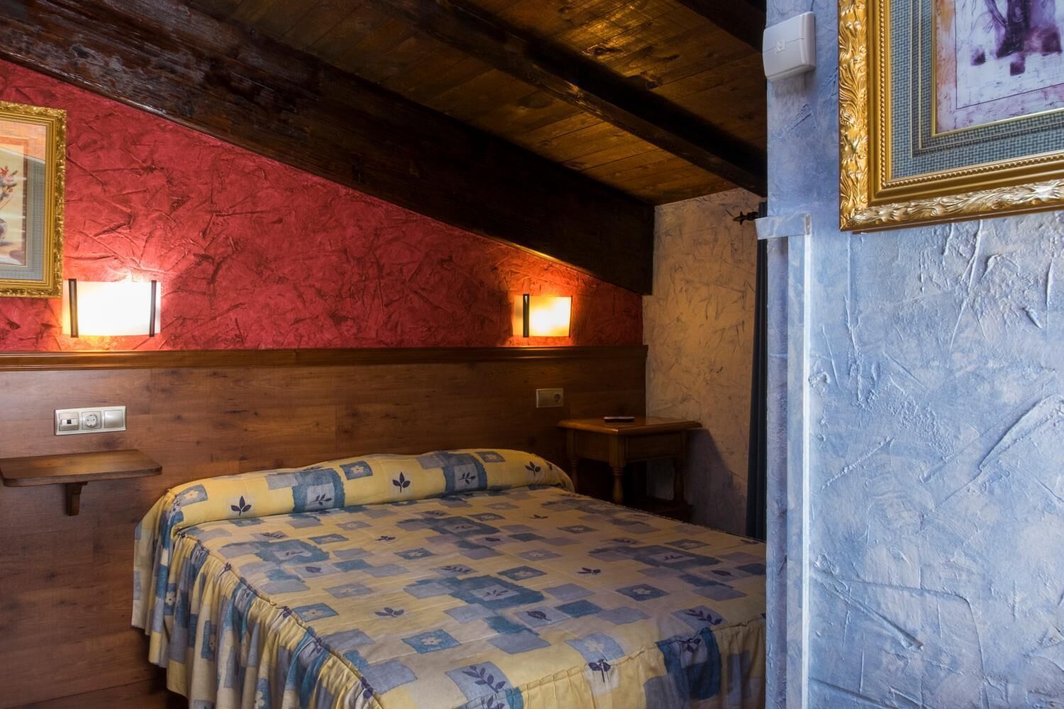 A cozy room with a wooden ceiling, a bed with a patterned bedspread, a small table, and two wall-mounted lights.