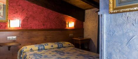 A cozy room with a wooden ceiling, a bed with a patterned bedspread, a small table, and two wall-mounted lights.