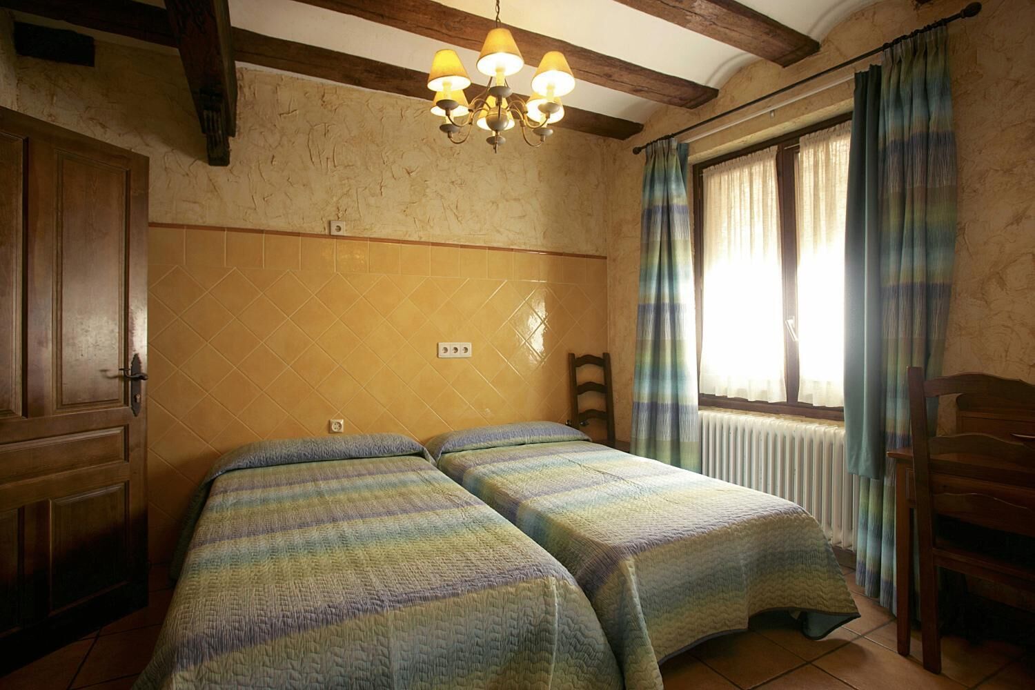 A room with two beds, wooden beams on the ceiling, a chandelier, a window with curtains, and a radiator.