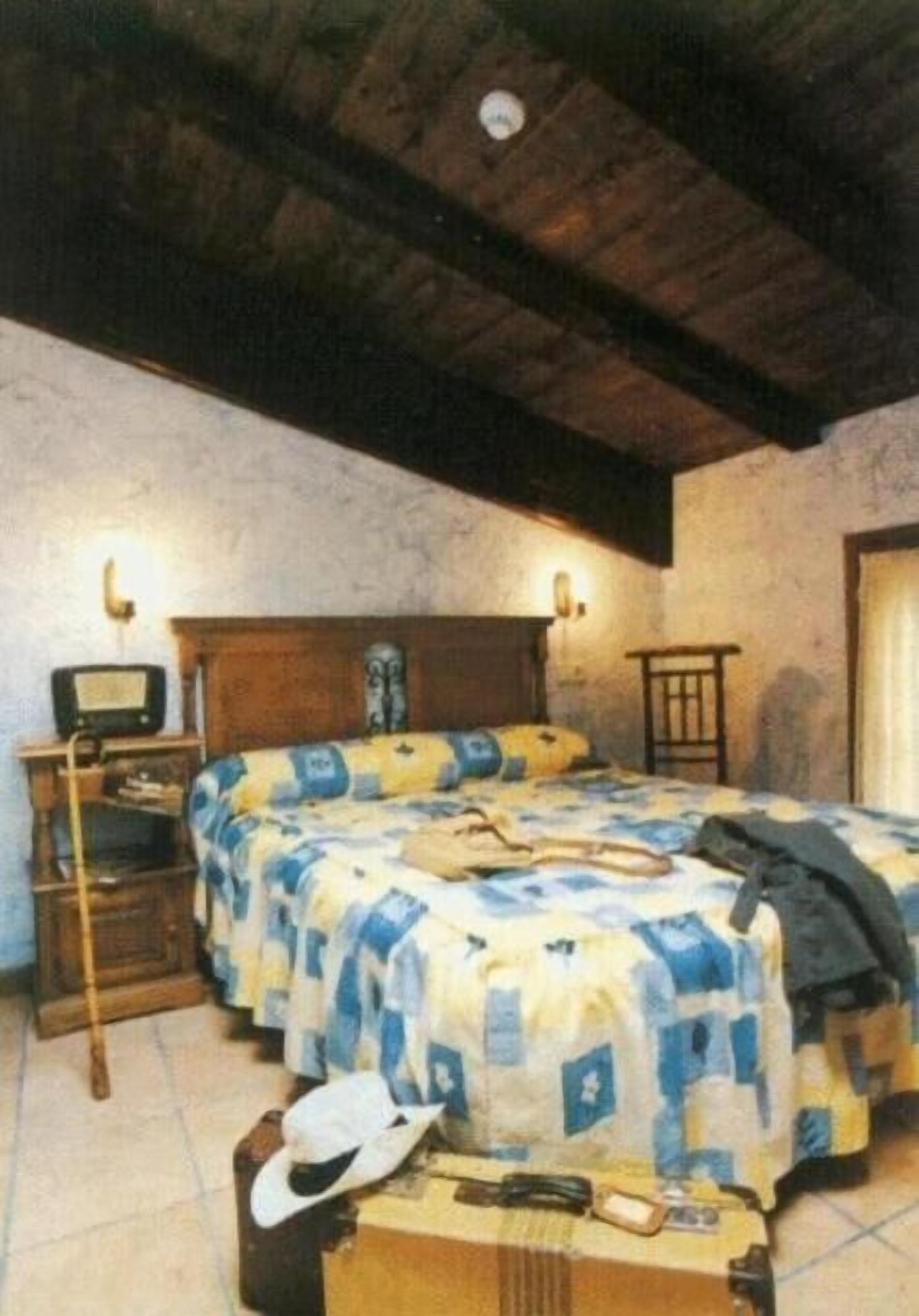 A bedroom with a wooden ceiling, a bed with a quilt, a bedside table with a lamp, and a suitcase on the floor.