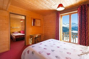 A wooden cabin room with a bed, a small table, a mirror, and a window with a view of snow-capped mountains.