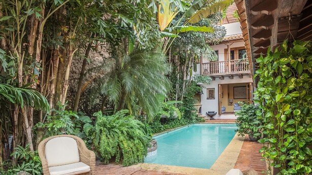 A lush indoor garden with a swimming pool, wicker furniture, and a building with a balcony. - Hotel Casa Mejía (Cartagena)