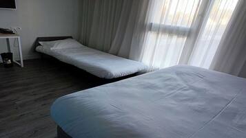 A hotel room with two single beds, a small desk with a telephone, and a window with sheer curtains.