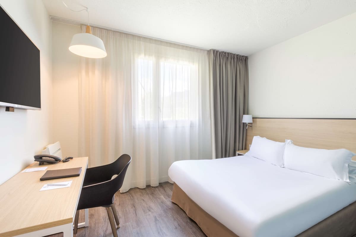 Een hotelkamer met een bed, een bureau met telefoon, een stoel, een aan de muur bevestigde televisie en een raam met gordijnen.
