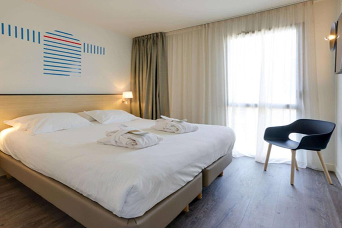 Une chambre d’hôtel avec un grand lit, deux lampes de chevet, une chaise et une fenêtre avec des rideaux.