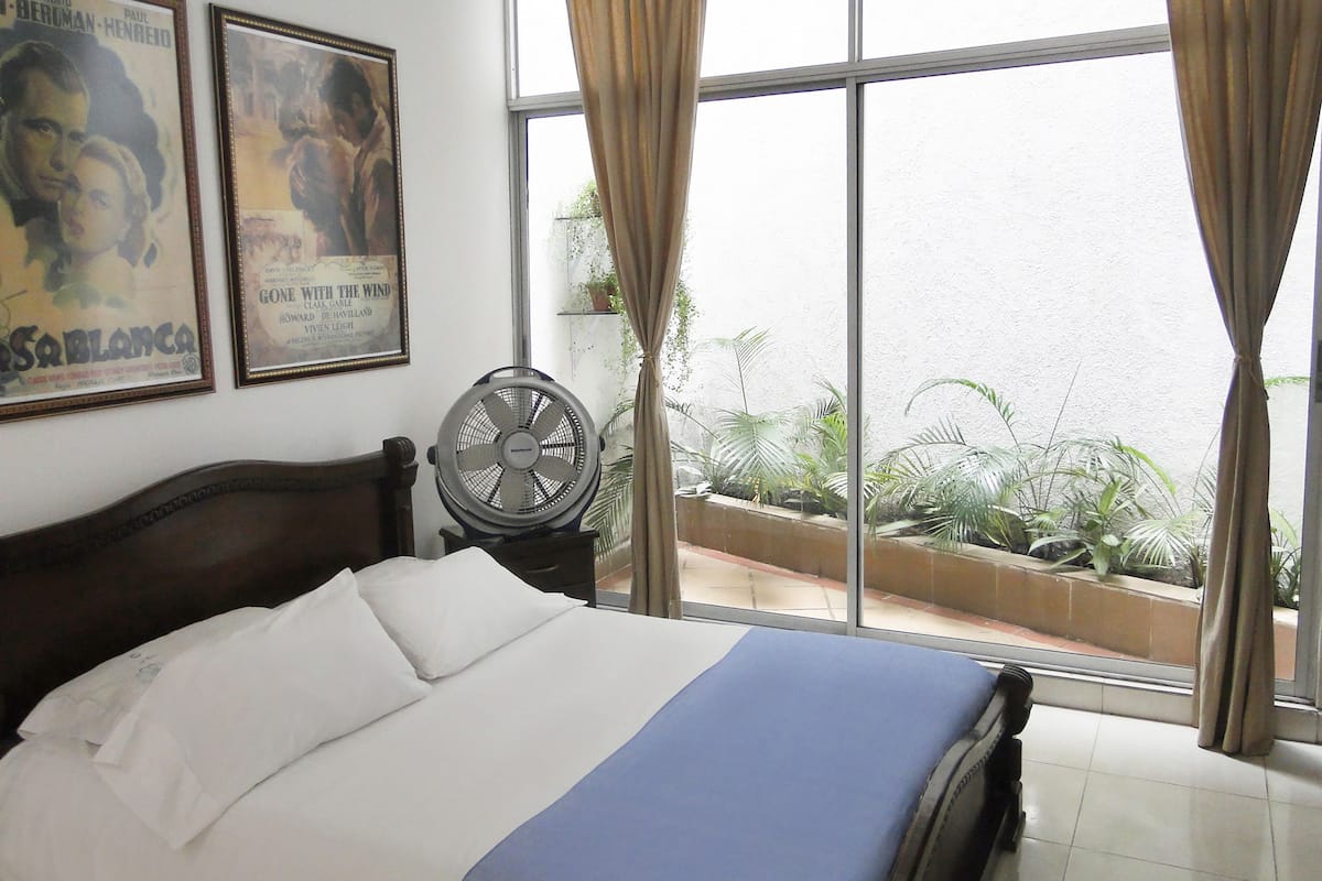 A hotel room with a bed, a standing fan, and large windows with curtains.