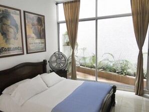 A hotel room with a bed, a standing fan, and large windows with curtains. - ARCO Apartasuites (Cali)