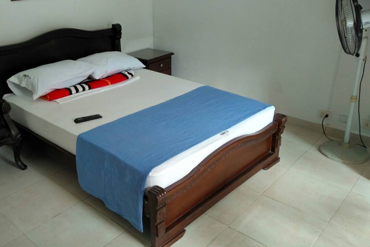 A single bed with a blue bedspread, a wooden headboard, a bedside table, a black chair, and a standing fan.
