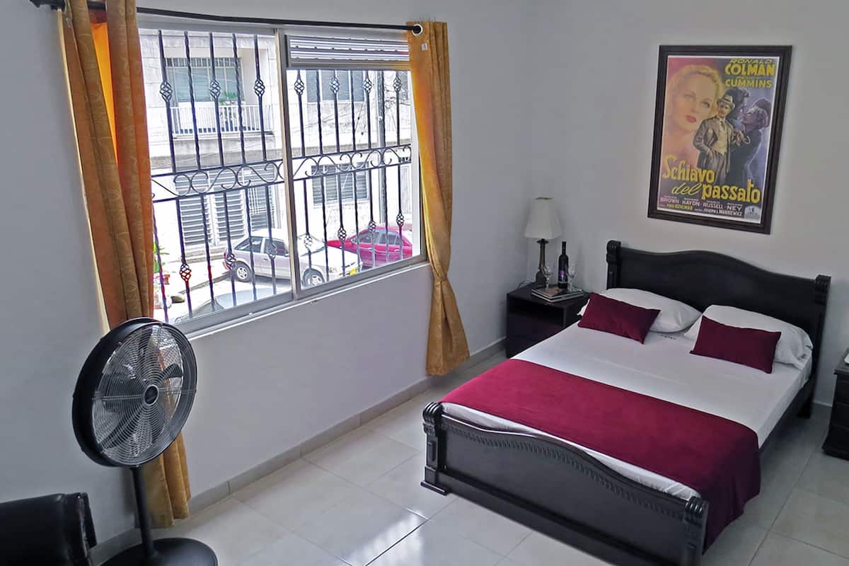 A bedroom with a bed, a nightstand, a lamp, a fan, a window with curtains, a poster on the wall, and a vase with flowers.