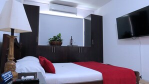 A hotel room with a bed, a TV mounted on the wall, a desk with a lamp, and a small plant on the dresser. - ARCO Apartasuites (Cali)