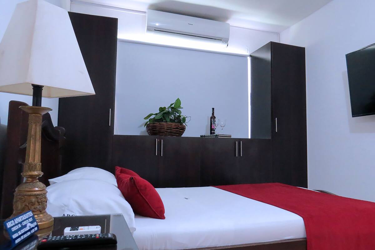 A hotel room with a bed, a TV mounted on the wall, a desk with a lamp, and a small plant on the dresser.