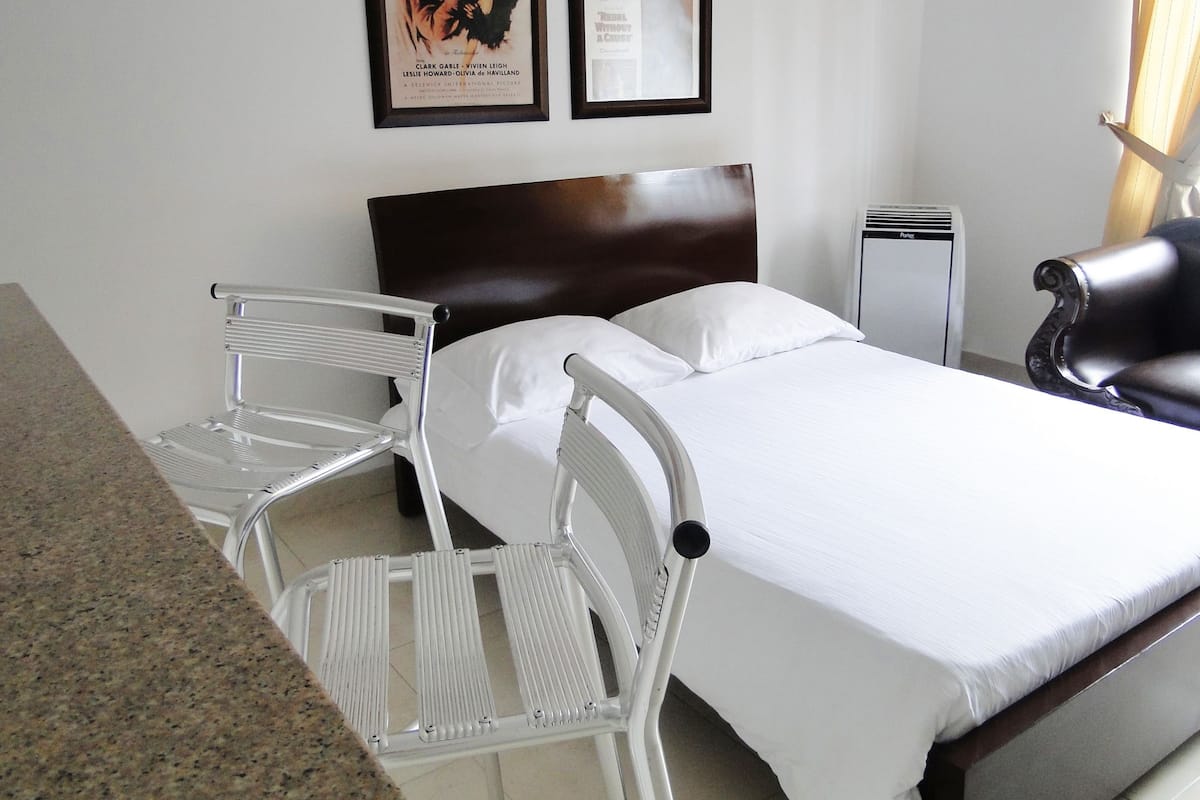 A hotel room with a bed, a white chair, a black chair, and two framed movie posters on the wall.