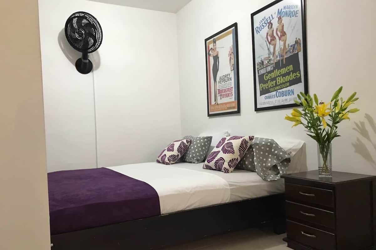 A bedroom with a bed, pillows, a bedside table, a fan, and a poster on the wall.