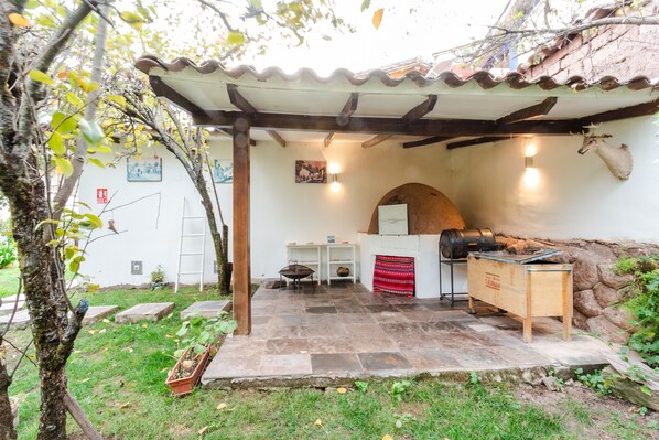 A covered outdoor patio with stone flooring, a wooden table, a grill, and a stone wall.
