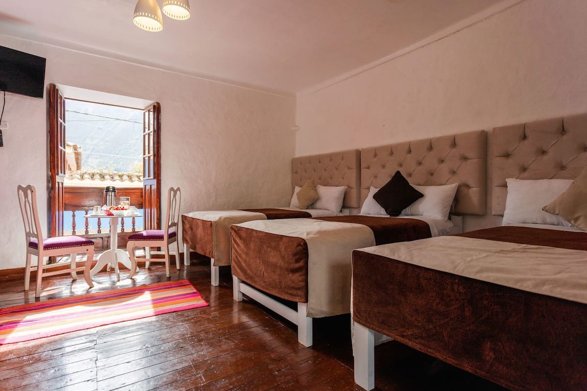 A hotel room with two beds, a dining area with a table and chairs, and a view through an open window.