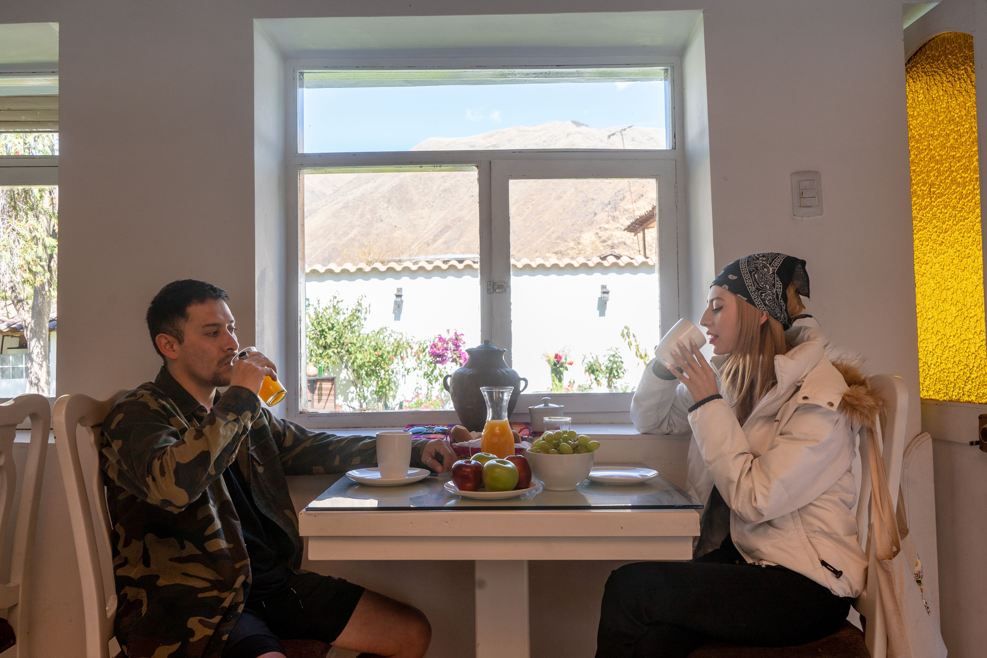 Two people seated at a table with breakfast items, a window with a view of a mountain and buildings, and a yellow textured wall.