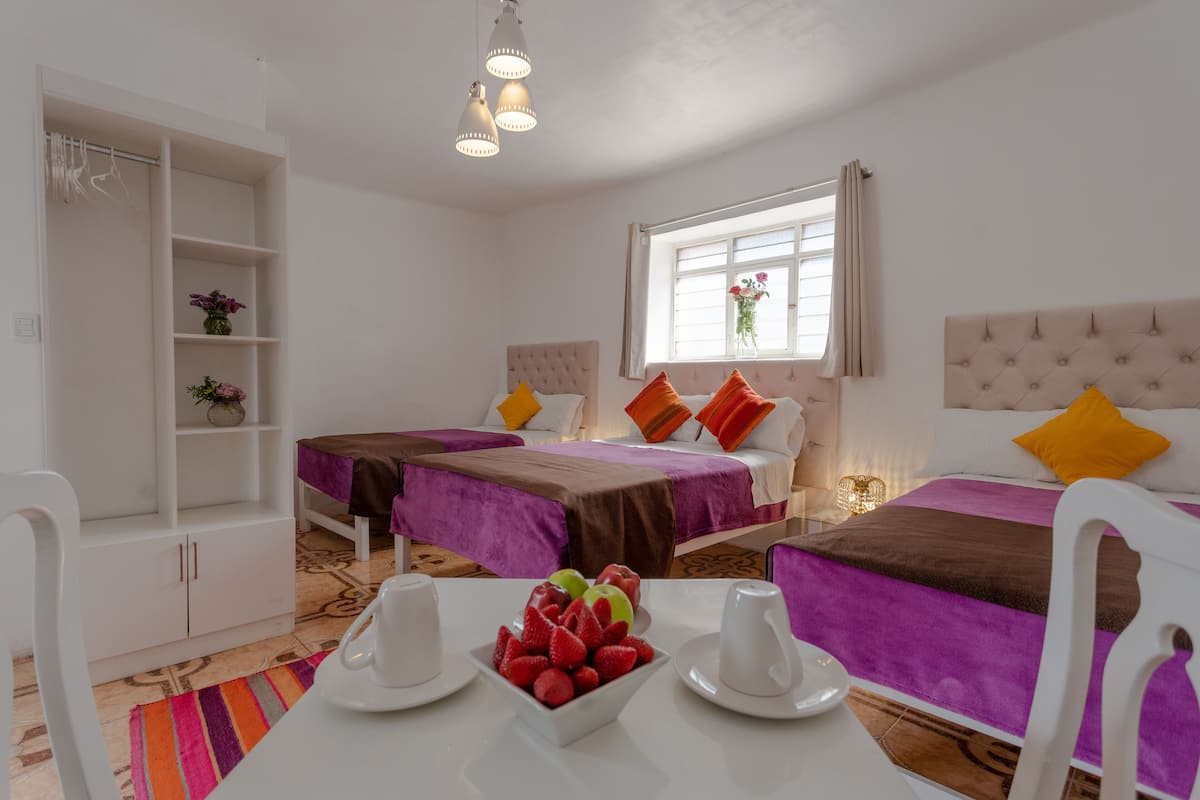 A hotel room with two beds, a dining table with fruit, a white wardrobe, and a window with flowers.