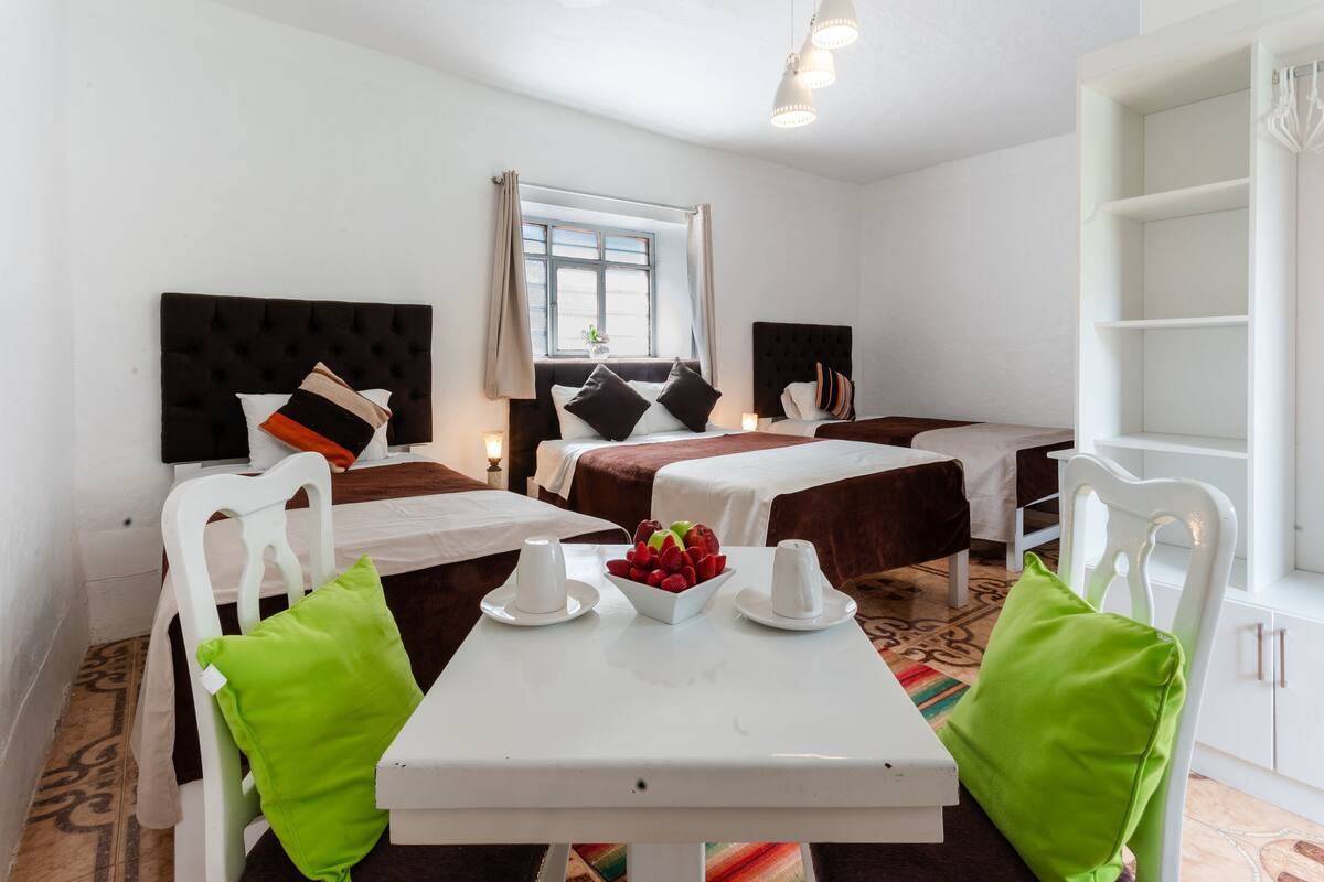A hotel room with a bed, two bedside tables, a window, and a dining area with chairs and a table.